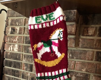 May include: A red, white, and green knitted Christmas stocking with the name "Eve" embroidered on the top. The stocking features a rocking horse with a pink body, yellow mane and tail, and a green saddle.