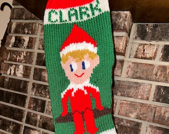 May include: A green knitted Christmas stocking with a red cuff and a white cuff. The stocking has a knitted image of an elf with a red hat, a white beard, and a brown belt. The stocking has the name "CLARK" knitted in white letters on the top.