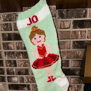 Puede incluir: Una media de Navidad de punto verde con un puño blanco y un diseño de bailarina roja y blanca. La media está personalizada con el nombre "JO".