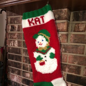 May include: A red, white and green knitted Christmas stocking with a snowman design and the name "KAT" embroidered on the front.