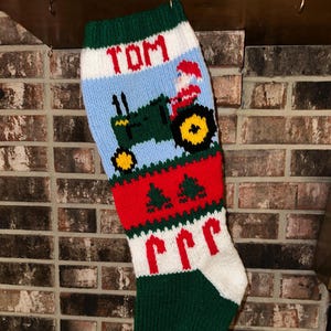 Könnte beinhalten: Handgestrickter Weihnachtsstrumpf in Grün, Rot, Weiß und Blau. Das Design zeigt einen Traktor mit Weihnachtsmann, den Namen "TOM" und Zuckerstangen. Spitze und Bündchen sind rot und grün, mit Backsteinhintergrund.