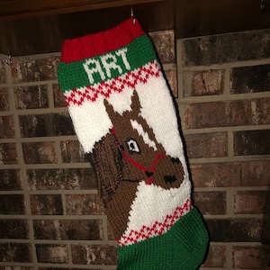 May include: A knitted Christmas stocking with a green, white, and red design. The stocking features a brown horse head with a red bridle and the name "Art" on the cuff.