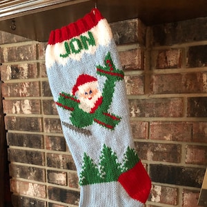 Könnte beinhalten: Ein gestrickter Weihnachtsstrumpf mit hellblauem Körper, roter Manschette und Spitze. Der Strumpf zeigt ein Weihnachtsmann-Design mit grünen Flügeln und dem Namen "JON" in weißen Buchstaben. Grüne Bäume sind ebenfalls abgebildet.