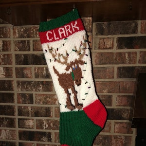 Peut inclure: Une chaussette de Noël blanche avec un revers vert et un bout rouge. La chaussette représente un renne brun avec un nœud papillon vert et une guirlande de lumières. La chaussette est personnalisée avec le nom "CLARK" en lettres rouges.
