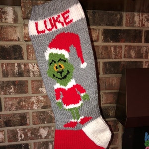 Peut inclure: Une chaussette de Noël en tricot gris et rouge avec un revers blanc. La chaussette représente un personnage vert du Grinch avec un bonnet de Père Noël rouge et le nom "LUKE" en lettres blanches.