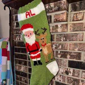 May include: A green knitted Christmas stocking with a white top and toe. The stocking features a Santa Claus and reindeer design. The initials "AL" are displayed in red on the white stripe.