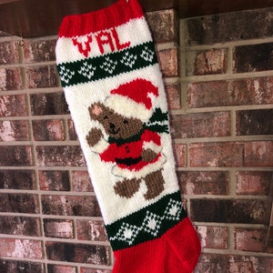 May include: A handmade Christmas stocking with a red cuff and toe, white body, and green and white accents. The stocking features a knitted teddy bear wearing a Santa hat and suit, with the name "VAL" knitted at the top.