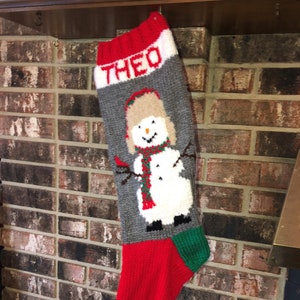 May include: Hand-knitted Christmas stocking with the name "THEO" in red letters. The stocking features a snowman design with a red scarf and a brown hat. The bottom is red with a green heel and toe.
