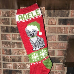 May include: Red and green knitted Christmas stocking with a white cat wearing a green Santa hat and the name "Adele" embroidered on the top.