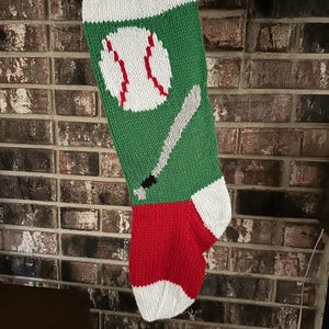 May include: A hand-knitted Christmas stocking featuring a green body with a white cuff, a red heel, and a white toe. The front displays a white baseball with red stitching and a grey baseball bat with a black handle, all created in knit yarn.