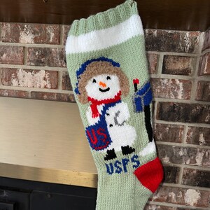 Op de afbeelding: Een groene gebreide kerstsok met een sneeuwpopontwerp. De sneeuwpop draagt ​​een blauwe muts, een rode sjaal en een blauw jasje. De sok heeft een witte streep en de woorden "USPS" in blauwe letters.