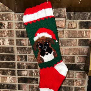 May include: Hand-knitted Christmas stocking featuring a Boxer dog wearing a Santa hat. The stocking is primarily green with red and white accents. The dog's face is brown, black, and white. The top and toe of the stocking are red.