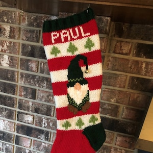 Puede incluir: Una media de Navidad de punto roja y verde con un diseño de gnomo y el nombre "PAUL" bordado en la parte superior. La media tiene una franja blanca con árboles de Navidad verdes.