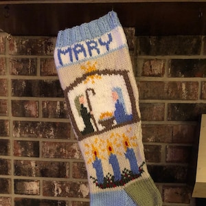 May include: A knitted Christmas stocking with the name "MARY" embroidered on the top. The stocking features a nativity scene with a brown wooden stable, a yellow star, and figures of Mary, Joseph, and a shepherd. The stocking is decorated with blue and white candles and green holly.