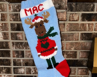 May include: A blue knitted Christmas stocking with a red and white trim. The stocking features a brown moose wearing a red sweater with a green and white scarf and a red Santa hat. The stocking is personalized with the name "MAC" in white letters.