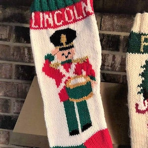 May include: A white knitted Christmas stocking with a red and green trim. The stocking features a cartoon toy soldier holding a drum and the name "LINCOLN" in red letters.