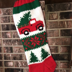 May include: Red and green knitted Christmas stocking featuring a red truck carrying a Christmas tree. The stocking has a white background with green and red accents.