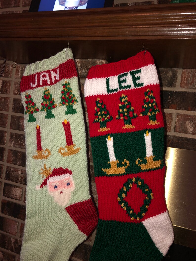 May include: Two knitted Christmas stockings with the names "Jan" and "Lee" on them. The stockings are decorated with green and red stripes, Christmas trees, candles, and a Santa Claus face.
