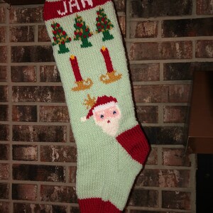 May include: A green knitted Christmas stocking with a red cuff and a white name "JAN" embroidered on the cuff. The stocking features a Santa Claus face, three Christmas trees, and three candles.