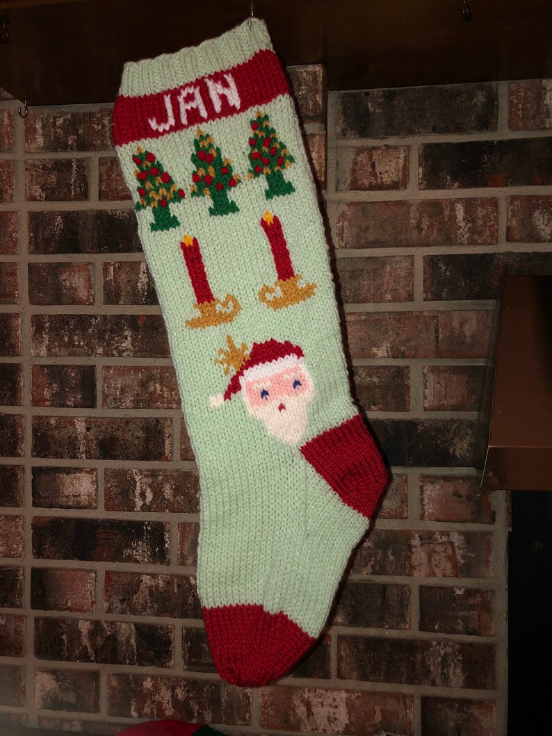 May include: A green knitted Christmas stocking with a red cuff and a white name "JAN" embroidered on the cuff. The stocking features a Santa Claus face, three Christmas trees, and three candles.