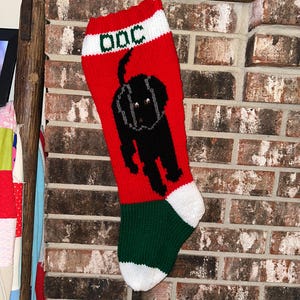 Könnte beinhalten: Ein rot-weiß-grüner Strick-Weihnachtsstrumpf mit einer schwarzen Hundegrafik auf der Vorderseite. Das weiße Bündchen oben zeigt den grünen Schriftzug "dog" in Blockschrift. Der Strumpf hat einen grünen Fersen- und Zehenbereich mit weißem Rand.