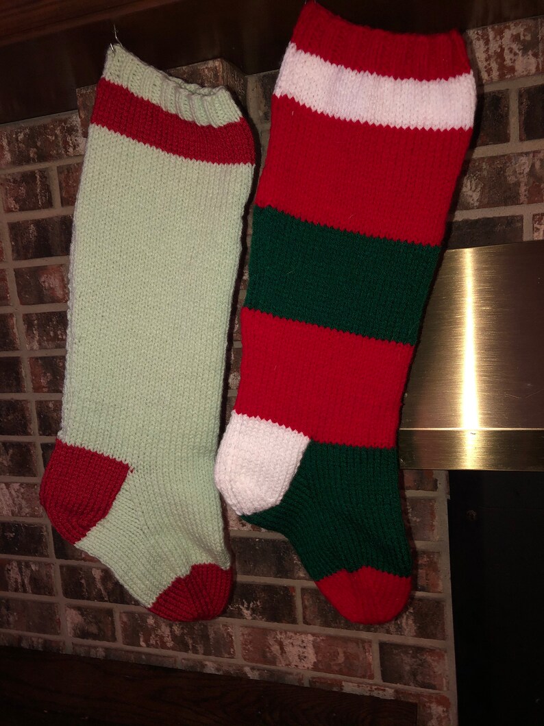 May include: Two knitted Christmas stockings hanging on a brick fireplace mantel. One stocking is green and white with red accents, and the other is red and green with white accents.