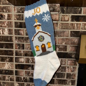 May include: Hand-knitted Christmas stocking with a church design. The stocking features a blue top with the name "JO" in yellow, a white snowflake, and a church with a cross. The bottom is white with a blue cuff.
