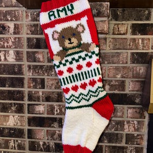 May include: A handmade Christmas stocking with the name "AMY" in green letters at the top. The stocking features a teddy bear design and red, white, and green accents. The stocking is hanging on a brick fireplace.