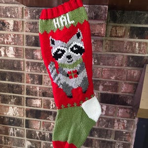 May include: A red and green knit Christmas stocking featuring a raccoon graphic holding a small red gift. The name HAL is knitted in white at the top. The stocking has a red cuff, a green heel, and a white toe section, all crafted in yarn.