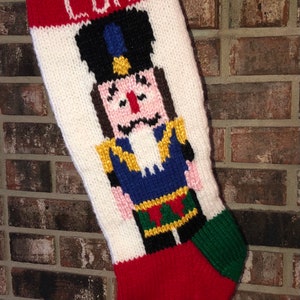 Puede incluir: Una media de Navidad de punto blanco con ribete rojo y verde. La media presenta una imagen tejida de un cascanueces con uniforme azul y sombrero dorado. La media está personalizada con el nombre "LUKE" en letras blancas.