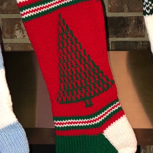 Könnte beinhalten: Ein rot-weiß-grüner gestrickter Weihnachtsstrumpf mit einem grünen Weihnachtsbaum-Motiv. Der Strumpf hängt an einem Kaminsims aus Ziegeln.