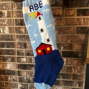 Op de afbeelding: Een gebreide kerstsok met een vuurtorenontwerp. De sok is voornamelijk lichtblauw met een witte vuurtoren, een rood gebouw en de naam "ABE" bovenaan. De vuurtoren heeft een gele straal.