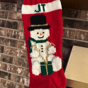 May include: Red knitted Christmas stocking featuring a snowman design. The snowman has a black top hat, green scarf, and gold detailing. The top of the stocking is white with the initials "JT" in green.