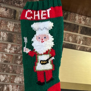 Peut inclure: Chaussette de Noël verte tricotée avec des détails rouges et le nom "CHET" en blanc. La chaussette présente un motif de chef Père Noël, tenant une spatule et une poêle. Le chef porte un chapeau et un tablier de chef blancs.