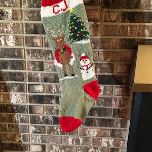 Peut inclure: Une chaussette de Noël tricotée à la main, vert sauge, avec un revers, un talon et une pointe rouges. Elle présente le nom CJ en lettres rouges, un renne brun avec une écharpe verte, un sapin de Noël décoré et un bonhomme de neige blanc.