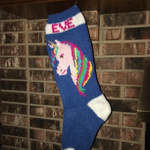 May include: A blue knitted Christmas stocking with a unicorn design. The unicorn has a pink face, a gold horn, and a rainbow mane. The word "EVE" is knitted in white letters at the top of the stocking.