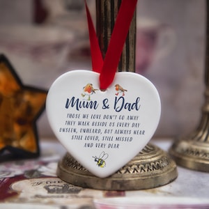 Könnte beinhalten: Weißer, herzförmiger Anhänger aus Keramik mit einem roten Band. Der Anhänger trägt eine blaue Inschrift mit dem Text: "Mama & Papa Die, die wir lieben, gehen nicht weg Sie gehen jeden Tag an unserer Seite Unsichtbar, unhörbar, aber immer nah Immer geliebt, immer vermisst und sehr lieb". Der Anhänger zeigt außerdem zwei kleine Abbildungen von Rotkehlchen und eine Abbildung einer Biene.