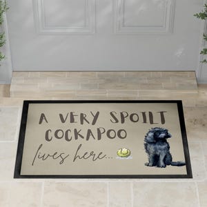Può includere: Zerbino rettangolare con bordo nero e sfondo beige. Il testo recita "A VERY SPOILT COCKAPOO lives here..." con un'illustrazione di un cane Cockapoo nero e una pallina da tennis.