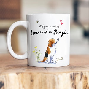 May include: White ceramic mug featuring a brown and white beagle illustration. The mug displays the text "All you need is Love and a Beagle" in black cursive writing. There is a small bee and two red hearts on the mug.