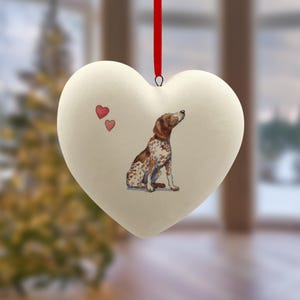 Op de afbeelding: Crèmekleurig hartvormig ornament met een aquarel schilderij van een hond. De hond is bruin en wit, omhoog kijkend. Twee kleine rode harten staan links. Een rood lint is bevestigd om op te hangen.