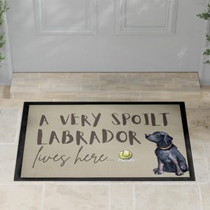 Op de afbeelding: Een rechthoekige deurmat met een zwarte rand en een beige achtergrond. De tekst "A VERY SPOILT LABRADOR lives here..." staat op de mat. Een zwarte Labrador-illustratie en een tennisbal zijn ook afgebeeld.