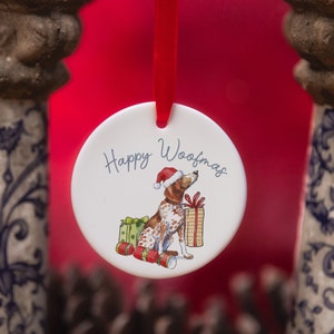 Op de afbeelding: Witte kerstbal van keramiek met een rood-witte hond die een kerstmanmuts draagt en een cadeau vasthoudt. De kerstbal heeft de tekst "Happy Woofmas".