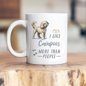 Könnte beinhalten: Weiße Keramik-Kaffeetasse mit einer Cartoon-Illustration eines Cavapoo-Hundes und dem Text "Ich mag Cavapoos mehr als Menschen".
