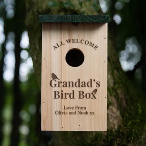 May include: A wooden birdhouse with a dark green roof is mounted on a tree. The birdhouse has a circular entrance hole and the text "ALL WELCOME" above it. Below the hole, the text reads "Grandad's Bird Box" with two small bird illustrations. The bottom of the birdhouse reads "Love From Olivia and Noah xx."