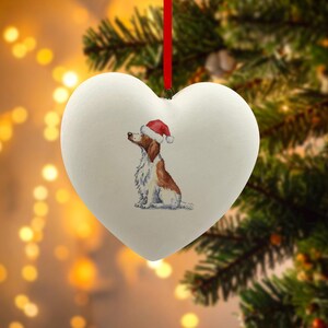 Puede incluir: Adorno navideño en forma de corazón blanco con una pintura de acuarela de un perro con un gorro de Papá Noel. El perro es marrón y blanco, con un gorro rojo y blanco. Cuelga de una cinta roja. Fondo con luces y ramas de pino.