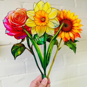 May include: A bouquet of three paper flowers, a red rose, a yellow daffodil, and an orange sunflower. The flowers are hand-painted and have a stained glass look.