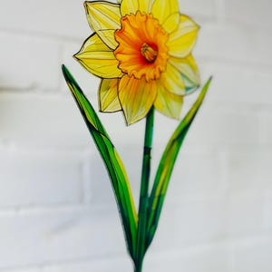 May include: A yellow daffodil flower with a green stem and leaves. The flower is made of stained glass and has a detailed design.