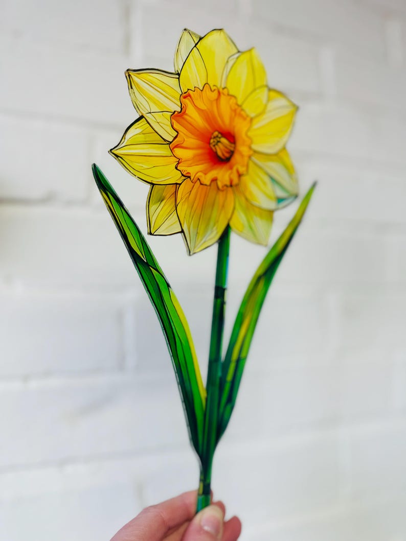May include: A yellow daffodil flower with a green stem and leaves. The flower is made of stained glass and has a detailed design.