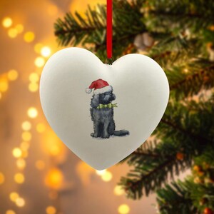 Könnte beinhalten: Herzförmiger Weihnachtsbaumschmuck mit einem gemalten schwarzen Hund, der einen Weihnachtsmannhut und eine grüne Fliege trägt. Der weiße Schmuck hängt an einem roten Band.