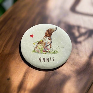 Può includere: Una pietra bianca con un'illustrazione di un cane marrone e bianco, circondato da fiori e un cuore rosso. Il nome "ANNIE" è scritto sotto il cane.